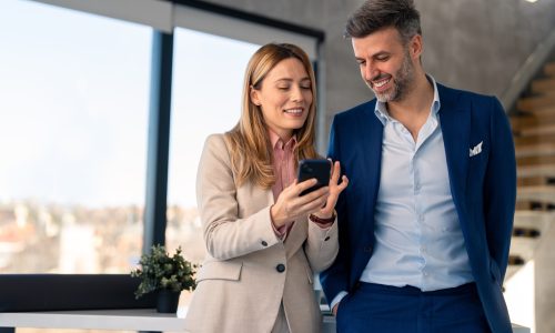 Young happy business woman wearing suit working in big modern city office with handsome coworker male CEO executive manager business man, using smart phone checking financial market data online.
